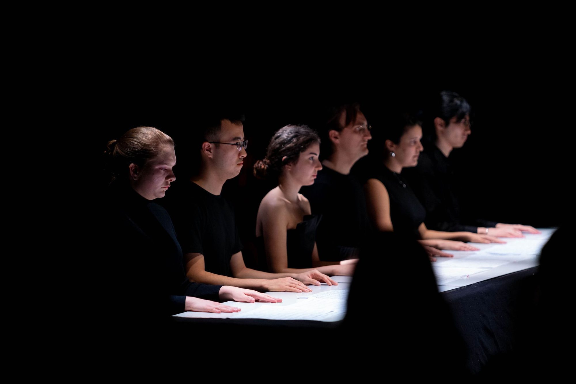 A row of performers with their hands on a lit table, IEMA
